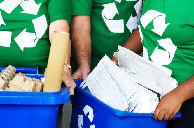 A imagem mostra três pessoas vestindo camisetas verdes com o símbolo de reciclagem em branco. Elas estão segurando duas caixas azuis de reciclagem. A caixa à esquerda contém materiais de papelão, incluindo um tubo de papelão e algumas caixas dobradas. A caixa à direita está cheia de papéis e documentos. As caixas azuis também têm o símbolo de reciclagem em branco. A cena sugere uma atividade de reciclagem de papel e papelão.