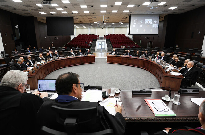 A imagem mostra uma sala de reuniões ampla, com um grande número de pessoas sentadas em torno de uma mesa em formato de "U". A sala tem um teto alto com várias luzes embutidas. No centro da mesa, há um espaço vazio, e as pessoas estão sentadas ao redor, olhando umas para as outras, sugerindo que estão participando de uma reunião ou discussão formal. Na parte de trás da sala, há fileiras de assentos vermelhos, semelhantes a um auditório, que estão vazios. Na parede do fundo, há duas grandes telas de projeção, uma das quais parece estar exibindo uma apresentação ou videoconferência. As pessoas na mesa estão vestidas formalmente, algumas usando togas, indicando que pode ser uma reunião de juízes ou um tribunal.