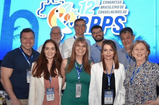 A imagem mostra um grupo de nove pessoas posando para uma foto em um evento. Ao fundo, há um painel azul com o texto "12º Congresso Brasileiro de Conselheiros de RPPS". As pessoas estão sorrindo e usando crachás pendurados em cordões azuis. A maioria está vestida de forma casual ou social, com camisas, blusas e jaquetas. O grupo é composto por homens e mulheres de diferentes idades.