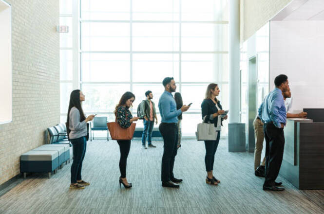 A imagem mostra um grupo de pessoas em fila dentro de um ambiente que parece ser um escritório ou uma agência. Há seis pessoas na fila, todas de pé, aguardando atendimento em um balcão à direita da imagem. A pessoa na frente da fila está interagindo com um atendente no balcão. As pessoas na fila estão vestidas de forma casual, algumas segurando celulares ou bolsas. O ambiente é iluminado por luz natural que entra através de grandes janelas ao fundo. Há também alguns assentos vazios encostados na parede à esquerda.