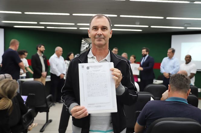 A imagem mostra um homem segurando um documento do programa Lar Legal. No fundo, há várias pessoas, algumas de pé e outras sentadas.
