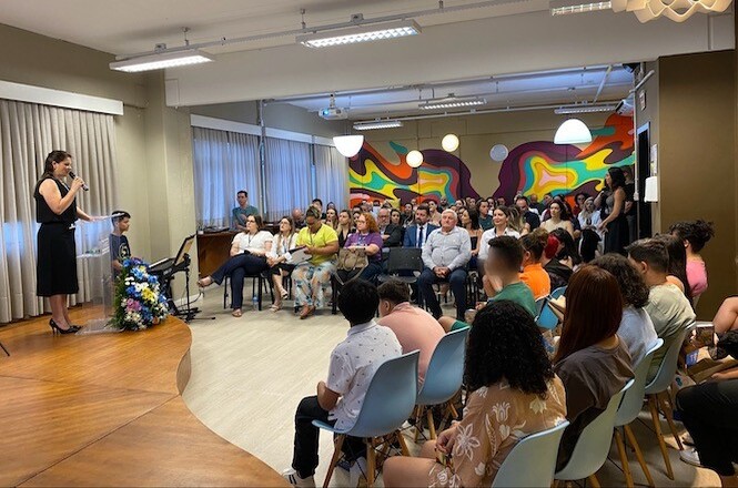 A imagem mostra um evento com os membros da Coordenadoria Estadual da Infância e da Juventude do Tribunal de Justiça de Santa Catarina e integrantes da Responsabilidade Social da FIESC. Uma mulher está falando em um palco, enquanto o público está sentado em cadeiras.
