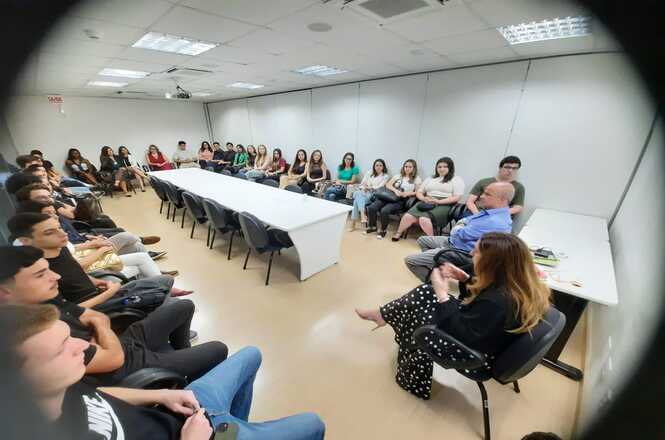 A imagem mostra uma turma de acadêmicos de Direito da Unisul, dispostos ao redor de uma mesa em formato retangular. Na parte da frente, estão o desembargador Júlio César Ferreira de Melo e a desembargadora Haidée Denise Grin, participando de uma conversa com os estudantes.