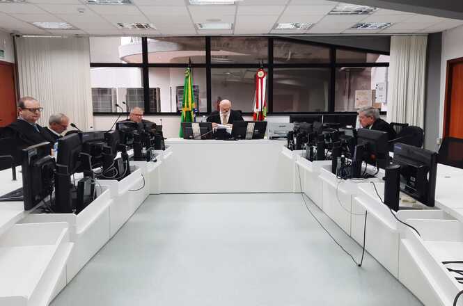 A imagem mostra uma sala na 4ª Câmara Comercial do Tribunal de Justiça, com pessoas sentadas ao redor de uma mesa em formato de "U". Há diversos computadores sobre a mesa e bandeiras do Brasil e do estado no fundo.