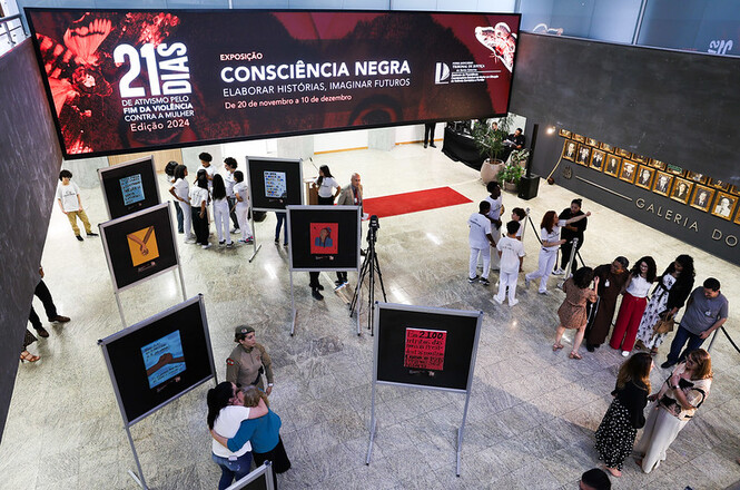 A imagem mostra a exposição de pinturas “Consciência Negra: Elaborar Histórias, Imaginar Futuros”. Pessoas estão circulando e olhando os quadros dispostos em cavaletes.