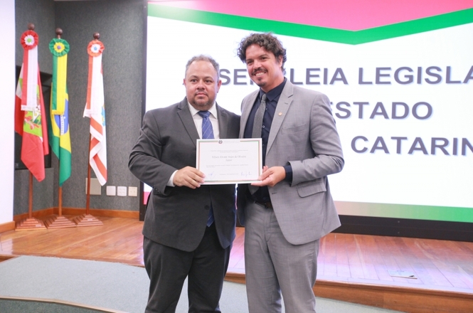 A imagem mostra o juiz Edison Alvanir Anjos de Oliveira Júnior e o deputado Marquito posando juntos para a foto. Os dois seguram um certificado. No fundo há  um painel com o texto "Assembleia Legislativa do Estado de Santa Catarina".