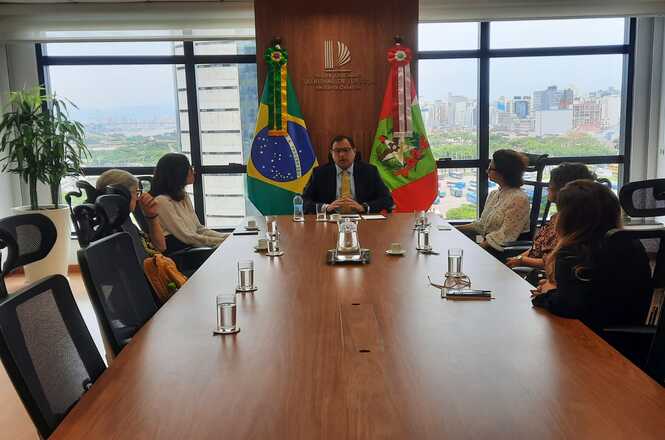 A imagem mostra uma sala de reunião com uma grande mesa de madeira, ao redor da qual estão sentados membros do Tribunal de Justiça, da Defensoria Pública Estadual e da Coordenadoria Nacional da Pastoral do Povo da Rua. Ao fundo, há bandeiras do Brasil e do estado.