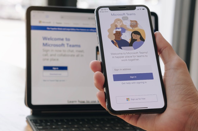 A imagem mostra uma pessoa segurando um smartphone que exibe a tela de login do Microsoft Teams. Ao fundo, há um laptop com a página inicial do mesmo aplicativo aberta, destacando o texto "Welcome to Microsoft Teams."
