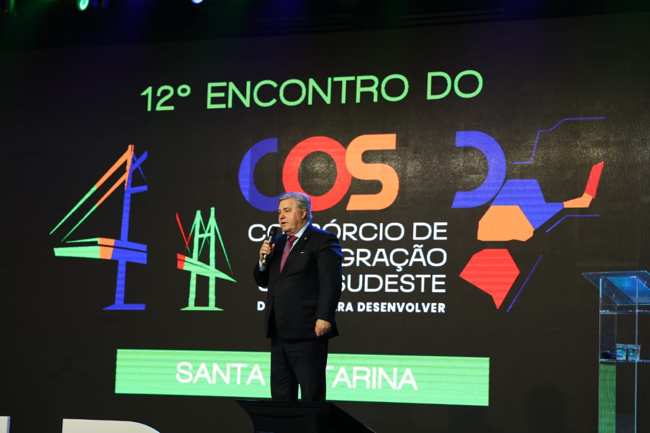 A imagem mostra o Desembargador Cid Goulart falando ao microfone em um palco durante o 12º Encontro do Consórcio de Integração Sul-Sudeste (COSUD). Ao fundo, há um telão com o logotipo do evento, incluindo ilustrações da ponte Hercílio Luz.
