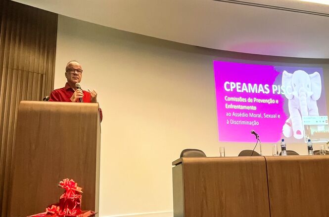 A imagem mostra um homem falando em um púlpito no evento “Ciclo de Debates: Assédio Moral e Sexual – Prevenção e Responsabilização Cível, Criminal e Administrativa.”. Na mesa ao lado do palestrante, estão dispostos microfones e garrafas de água.