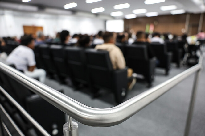 A imagem mostra um auditório com cadeiras pretas, onde várias pessoas estão sentadas. O foco da foto está em um corrimão metálico na parte frontal, enquanto o fundo, onde ocorre o evento, está desfocado.