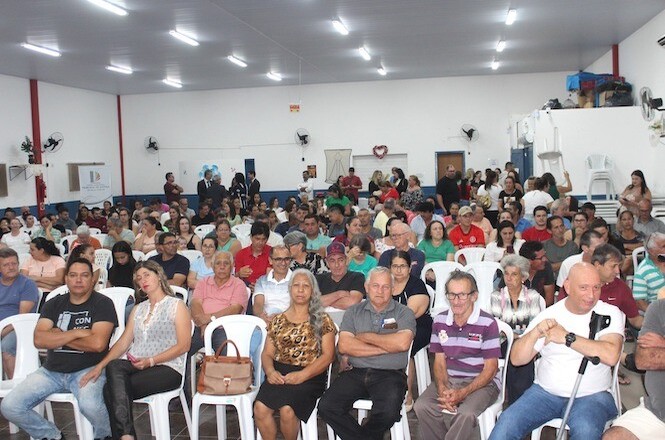 A imagem mostra uma fotografia de um grupo numeroso de pessoas sentadas em cadeiras brancas em um salão amplo, assistindo a um evento. As cores predominantes do local são tons de cinza, branco e vermelho.