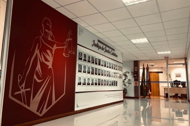 A imagem mostra o interior de um prédio institucional À esquerda, há uma grande parede vermelha com a figura da deusa da Justiça, ao lado de outra parede branca que exibe quadros com fotos de pessoas, provavelmente homenagens a profissionais ou autoridades. Ao fundo, há portas de vidro, bandeiras oficiais e plantas decorativas, reforçando o contexto formal do espaço.