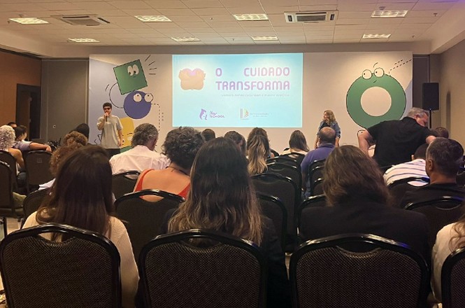 A imagem mostra um auditório iluminado, com pessoas sentadas em cadeiras pretas, assistindo a uma apresentação projetada em tela grande com o tema "O cuidado transforma". As paredes exibem formas coloridas em verde, roxo e laranja, complementando o design do slide.
