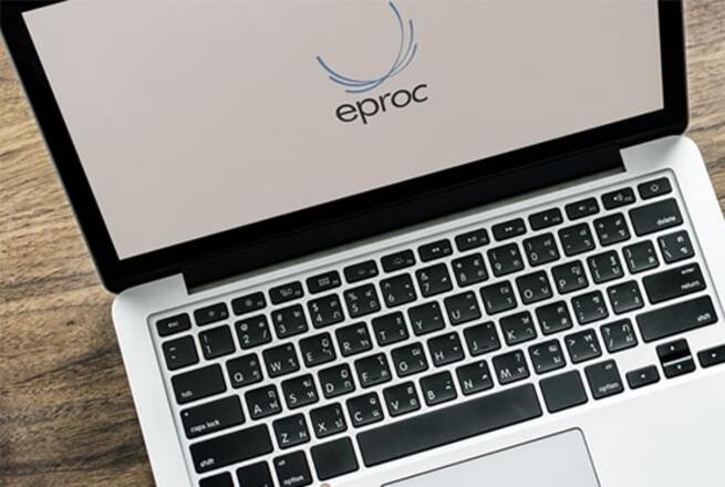 A imagem mostra um notebook sobre uma mesa de madeira. Na tela do notebook, está exibido o logotipo do sistema "eproc". O design do aparelho é moderno.