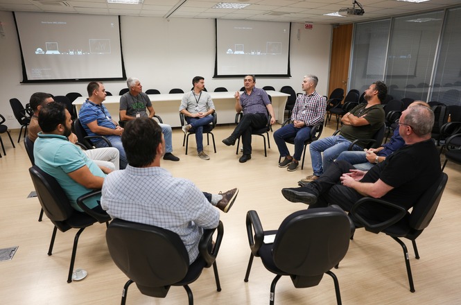A imagem mostra um grupo de homens sentados em um círculo dentro de uma sala de reuniões. O ambiente possui paredes brancas, um piso de madeira clara e cadeiras pretas. No fundo, há duas telas de projeção fixadas na parede.