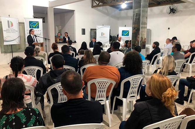 A imagem mostra a entrega dos títulos de propriedades do Lar Legal em espaço interno, onde pessoas estão sentadas em cadeiras plásticas brancas, voltadas para o desembargador Selso de Oliveira que está de pé ao lado de um púlpito. No fundo estão banners identificando o Tribunal de Justiça.