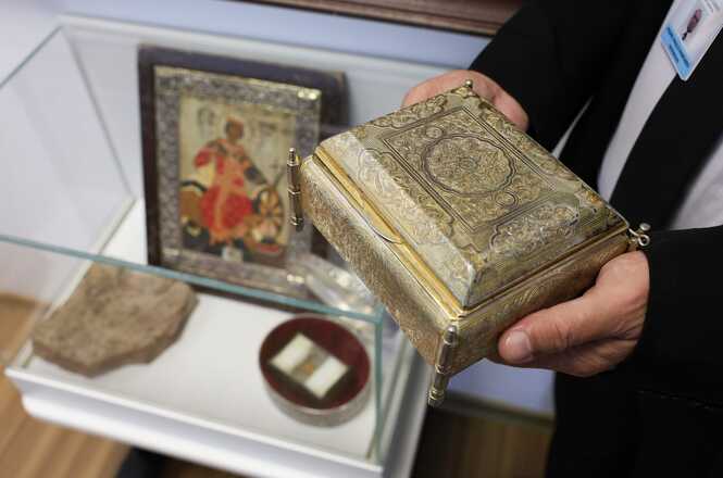 A imagem mostra mãos que seguram um relicário dourado com detalhes gravados. Ao fundo, uma vitrine exibe outras relíquias de Santa Catarina de Alexandria, incluindo um quadro com moldura marrom.