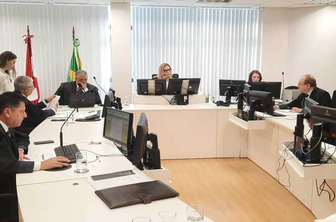 A imagem mostra uma sala de audiência clara, com móveis brancos e piso de madeira, onde a desembargadora Maria do Rocio Luz Santa Ritta e outros profissionais do Judiciário estão sentados.