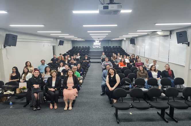 A imagem mostra um auditório, com cadeiras pretas organizadas em fileiras, com diversas pessoas, posando para a foto na palestra da desembargadora Hildemar Meneguzzi de Carvalho. O ambiente é amplo e tem o teto branco.