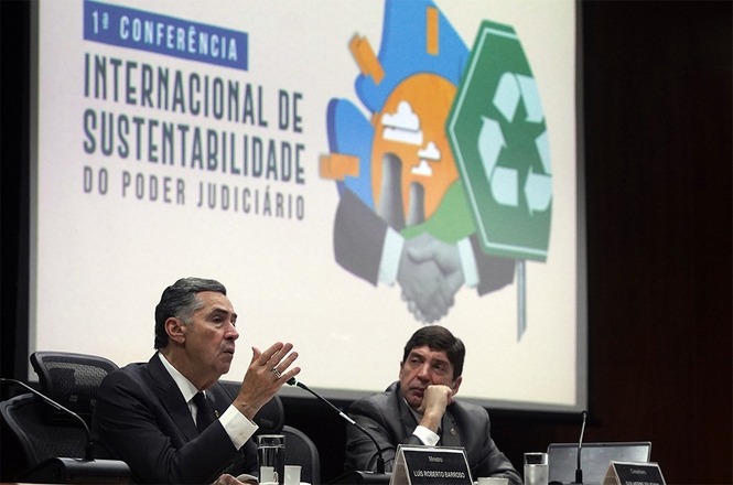 A imagem mostra dois homens participando de uma mesa durante a 1ª Conferência Internacional de Sustentabilidade do Poder Judiciário. À esquerda, um dos palestrantes, usando terno preto e gravata escura, gesticula enquanto fala. À direita, outro participante, também de terno, apoia o rosto na mão.