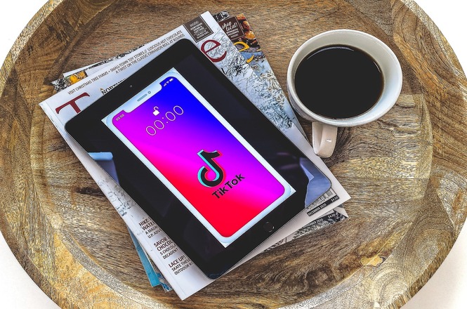 A imagem mostra um tablet com a tela inicial aberta no aplicativo Tik Tok colocado em uma bandeja de madeira. Ao lado, há uma xícara de café preto e algumas revistas sob o tablet, incluindo uma edição da revista Time.