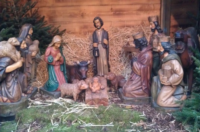 A imagem mostra uma representação de um presépio tradicional, com figuras de madeira esculpidas e pintadas. Mostra Maria, José, o Menino Jesus em uma manjedoura, junto com animais (ovelhas, um boi), os Três Reis Magos e outros personagens. O cenário está decorado com palha e árvores ao fundo, criando um ambiente rústico e natalino.