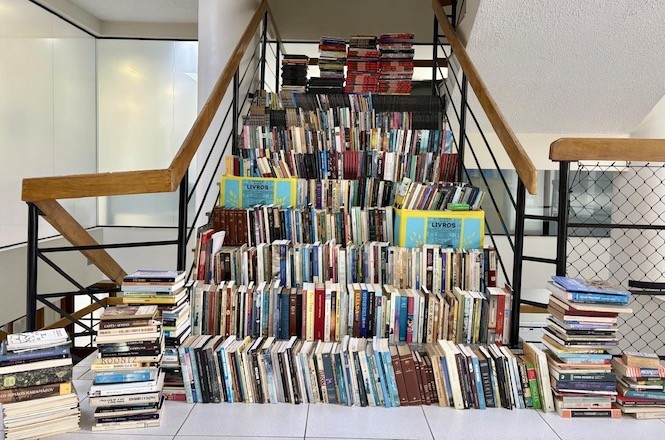 A imagem mostra uma escadaria completamente decorada com livros organizados em uma estrutura que forma um "mural" de livros, cobrindo os degraus. Há pilhas de livros adicionais ao lado da escada, e algumas caixas identificadas com a palavra "LIVROS". O ambiente é interno, iluminado, e parece ser um espaço público ou institucional, como uma biblioteca ou um local para troca ou doação de livros. A organização transmite uma sensação de incentivo à leitura.