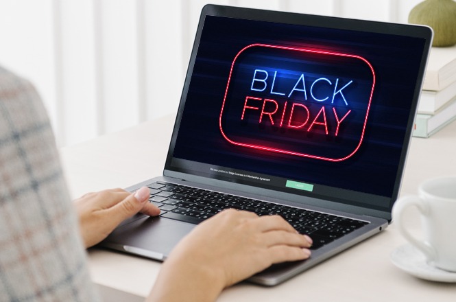 A imagem mostra uma pessoa usando um laptop com um letreiro de "Black Friday" em neon na tela. Na mesa clara, há uma xícara e livros, sugerindo um ambiente organizado e tranquilo.