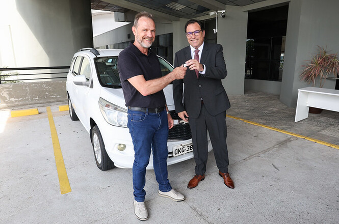 A imagem mostra um veículo, Chevrolet Spin. Em frente ao carro, o desembargador Francisco Oliveira Neto entrega a chave do automóvel ao gerente administrativo da unidade hospitalar, Nélio Francisco Schmitt.