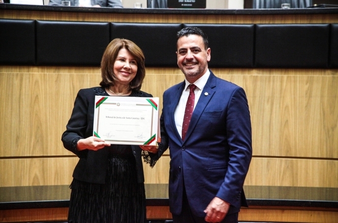 A imagem mostra a desembargadora Janice Goulart Garcia Ubialli, usando roupas pretas e segurando um certificado. Do seu lado, um homem vestido com terno azul, entrega o documento.
