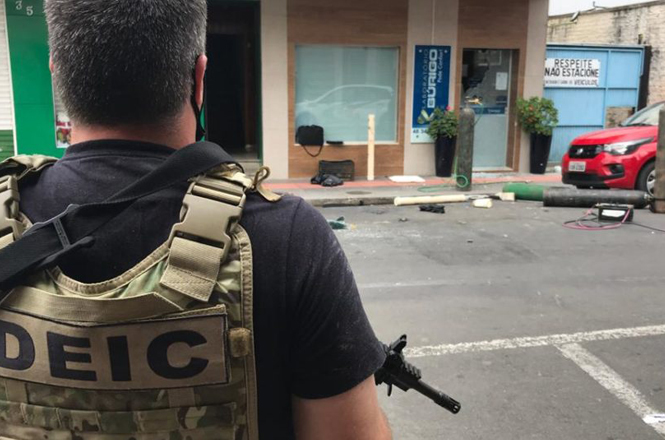 A imagem mostra um policial de costas, ele segura uma arma. No fundo tem algumas lojas e um carro vermelho.