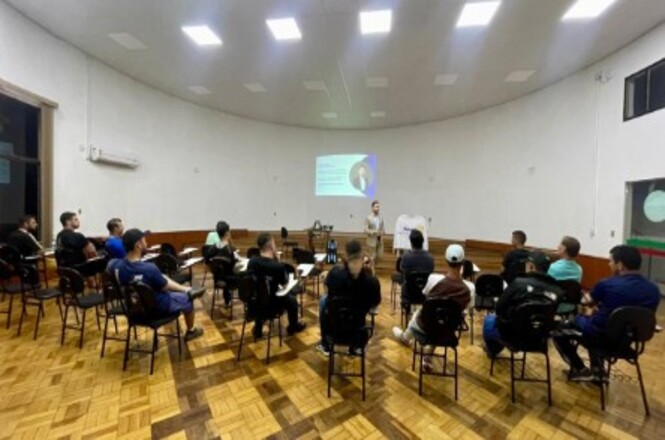A imagem mostra vários homens sentados em carteiras escolares em uma sala redonda. O chão é de madeira e as paredes são brancas. No fundo, um homem está palestrando, com uma projeção na parede.