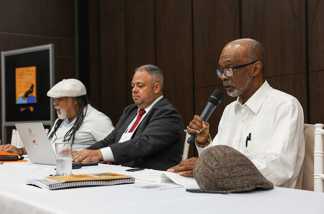 A imagem retrata três homens sentados em uma mesa no seminário “Questões Étnico-Raciais na Perspectiva da Interseccionalidade de Gênero e Raça”. O homem que está com o microfone palestrando usa roupa branca. Em cima da mesa tem livros, papeis, copos de água e um laptop.