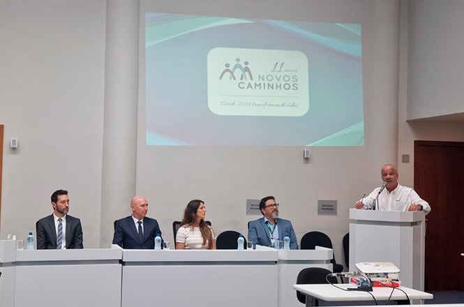 A imagem mostra uma mesa composta por quatro pessoas sentadas e ao lado o desembargador Júlio César Ferreira de Melo em um púlpito. Ele está de camisa branca, gesticulando enquanto fala ao público. Ao fundo, há uma projeção com o logotipo do programa "Novos Caminhos".