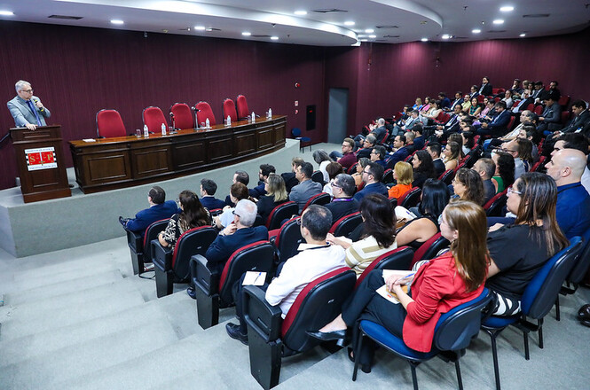 A imagem mostra um auditório. Muitas pessoas estão sentadas na plateia enquanto um homem de terno cinza fala em um púlpito no palco.