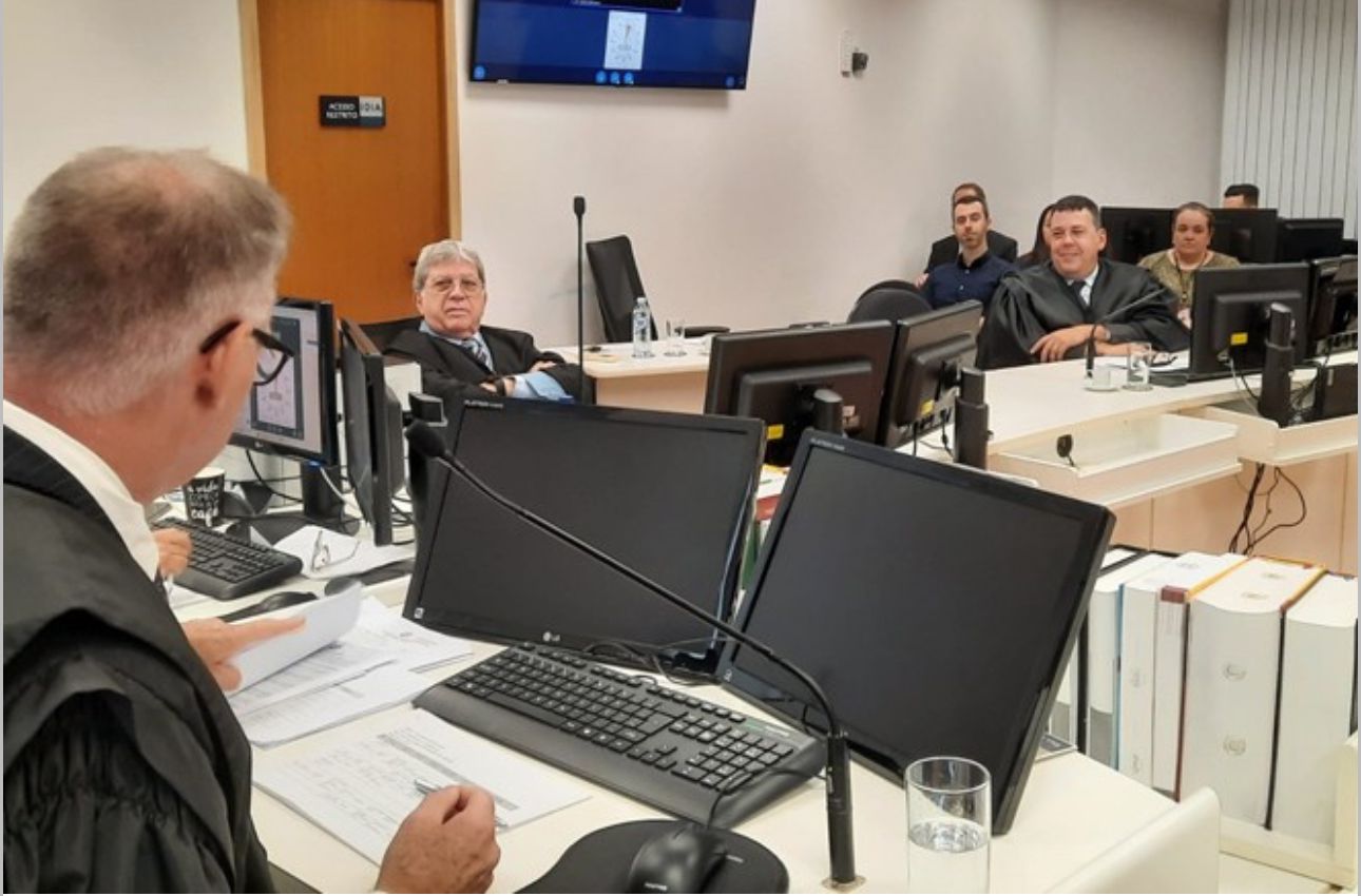A imagem mostra um sala com pessoas sentadas em mesas com computadores e documentos, dentre elas está o desembargador Sebastião César Evangelista. Todos estão vestindo trajes judiciais. Os rostos estão obscurecidos para privacidade.