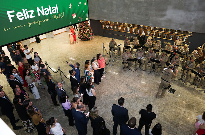 A imagem mostra uma cerimônia ocorrendo no hall de entrada do TJSC, com várias pessoas em pé observando uma apresentação musical feita por uma banda composta por integrantes da Polícia Militar. Ao fundo, um grande painel eletrônico exibe a mensagem "Feliz Natal e um próspero 2025!". Há também uma árvore de Natal decorada no canto.