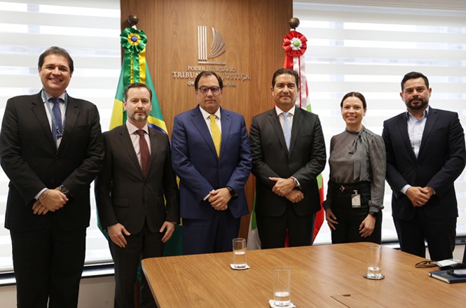 A imagem mostra um grupo de seis pessoas em um ambiente formal, entre elas o presidente do Tribunal de Justiça de Santa Catarina (TJSC), desembargador Francisco Oliveira Neto. Há uma bandeira do Brasil, de Santa Catarina e uma placa com o emblema do Tribunal de Contas da União (TCU) na parede atrás deles. Há, também, uma mesa à frente deles.