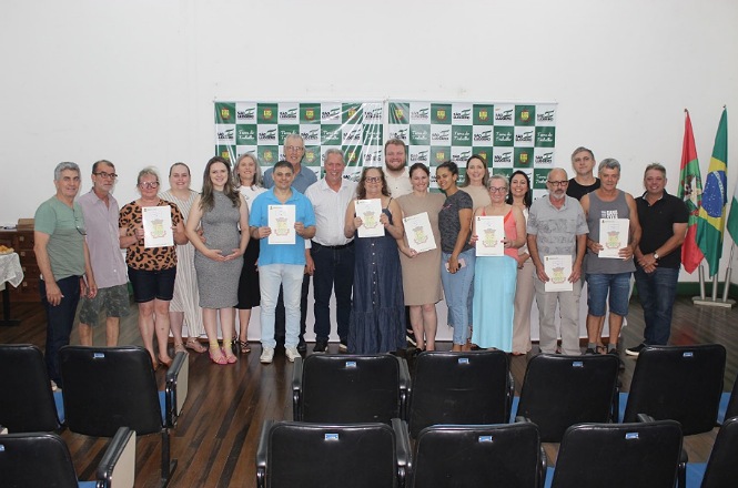 Esta imagem mostra um grupo de pessoas posando para uma foto em uma sala. Todos estão alinhados em duas fileiras. Algumas estão segurando certificados ou documentos com capas ilustradas. Atrás do grupo, há um painel com logotipos e, no lado direito, aparecem as bandeiras do Brasil e de Santa Catarina.