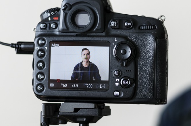 A imagem mostra a tela de uma câmera digital. Na tela, é visível a imagem de um homem em um fundo branco sendo filmado. A câmera está montada em um tripé, e os botões são visíveis.