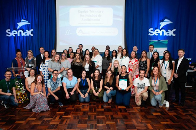 A imagem mostra um grupo grande de pessoas posicionado para uma foto em frente a um fundo azul com o logo do Senac ao lado. Todos estão sorrindo, e atrás há uma tela de projeção exibindo informações sobre um evento.