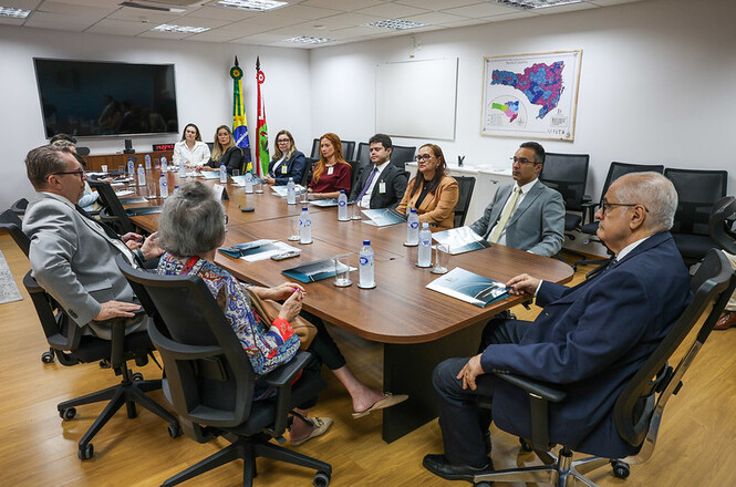 A imagem mostra nove pessoas em uma sala de reunião formal, sentadas ao redor de uma mesa com garrafas de água e documentos. No fundo, há bandeiras, um mapa na parede e uma TV. O ambiente é claro e organizado.