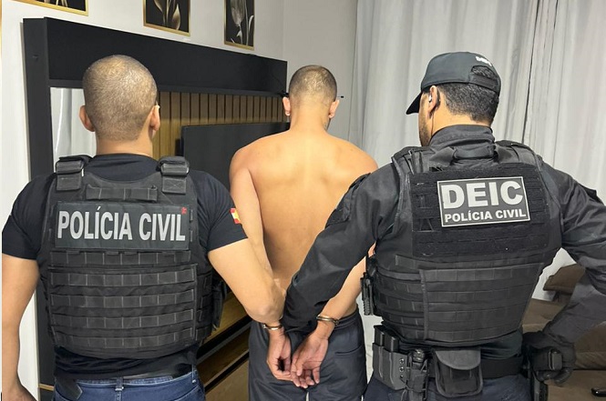 A imagem mostra dois agentes da Polícia Civil conduzindo um homem algemado. O homem está sem camisa, de costas e com as mãos presas por algemas. Os agentes estão usando coletes pretos, sendo que um deles possui a inscrição "POLÍCIA CIVIL" nas costas e o outro, "DEIC POLÍCIA CIVIL".
