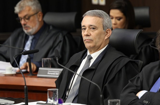 A imagem mostra o magistrado Carlos Roberto da Silva, vestido com uma toga em um sessão no pleno do TJSC. Ele está sentado, na mesa em sua frente tem microfone, copos de água e papeis.
