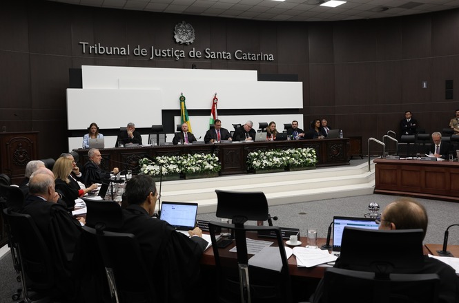 A imagem mostra uma sessão em andamento no plenário do Tribunal de Justiça de Santa Catarina. Magistrados e servidores estão sentados em mesas organizadas em formato semicircular. O espaço é decorado com flores brancas e possui o brasão do tribunal ao fundo.