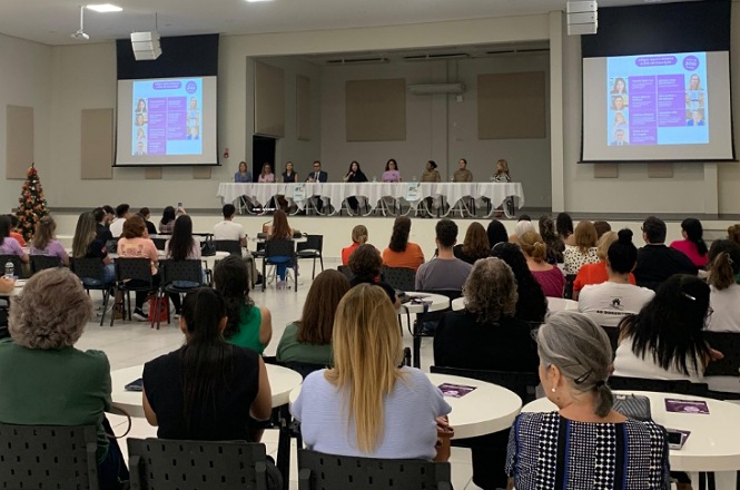 A imagem mostra um auditório onde ocorre o evento “Lei Maria da Penha em Debate”. No palco, há uma mesa composta por várias palestrantes, com microfones e telas grandes projetando informações. O público está sentado em mesas redondas.