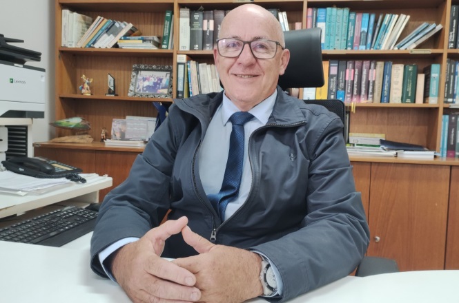 A imagem mostra o desembargador Antônio Carlos Junckes dos Santos sentado, vestindo camisa, gravata e uma jaqueta Ao fundo, há uma estante com diversos livros e sobre a mesa, é possível ver um telefone, documentos e outros itens de trabalho.