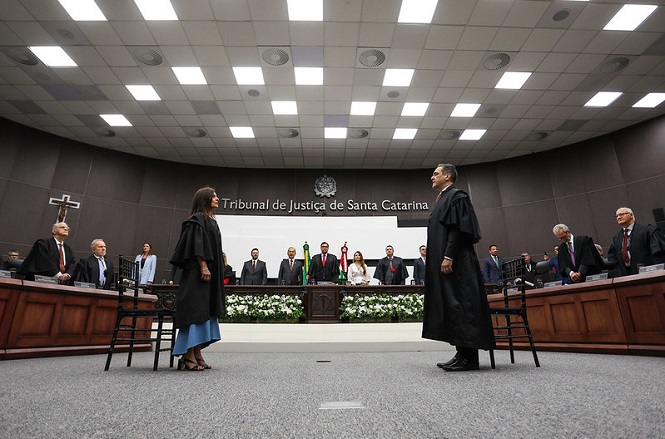 A imagem mostra uma cerimônia de posse no Tribunal de Justiça de Santa Catarina, com duas pessoas em posição de destaque no centro, trajando togas. Ao fundo, estão outras autoridades, com uma mesa decorada com arranjos de flores.