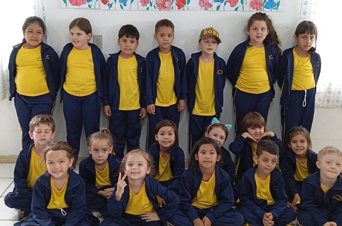 A imagem mostra um grupo de crianças posado para uma foto. Elas vestem uniformes escolares compostos por camisetas amarelas e jaquetas e calças azuis. Ao fundo, há uma parede decorada com desenhos coloridos.